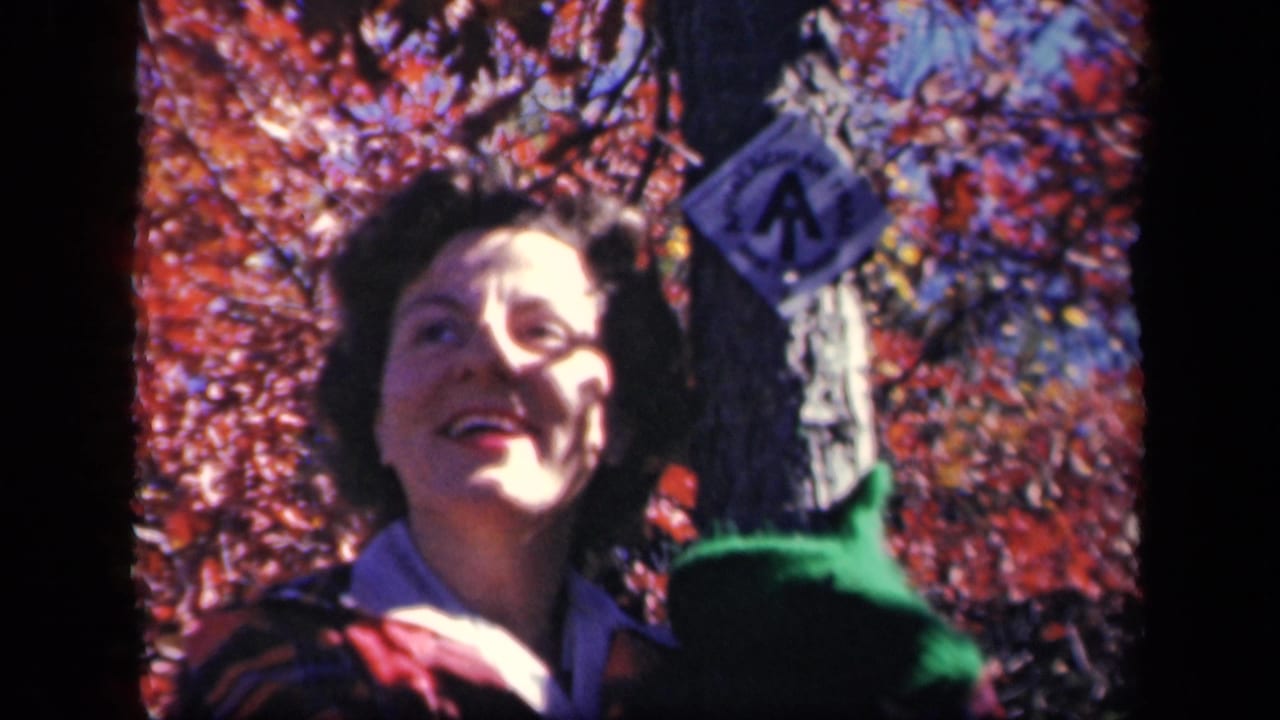 A woman smiles toward the camera near a trail marker at Skyline Drive, Virginia, with red autumn leaves and sunlight across her face.