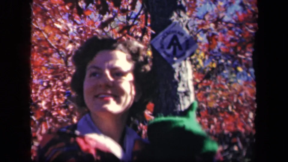 A 1940s home-movie frame shows a woman smiling near a tree and trail marker, with red leaves filling the background.