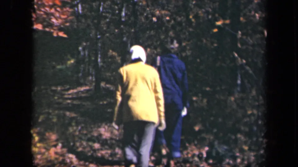 Two women continue away from the camera on a leaf-strewn forest path, with one wearing a yellow jacket and the other dressed in blue.