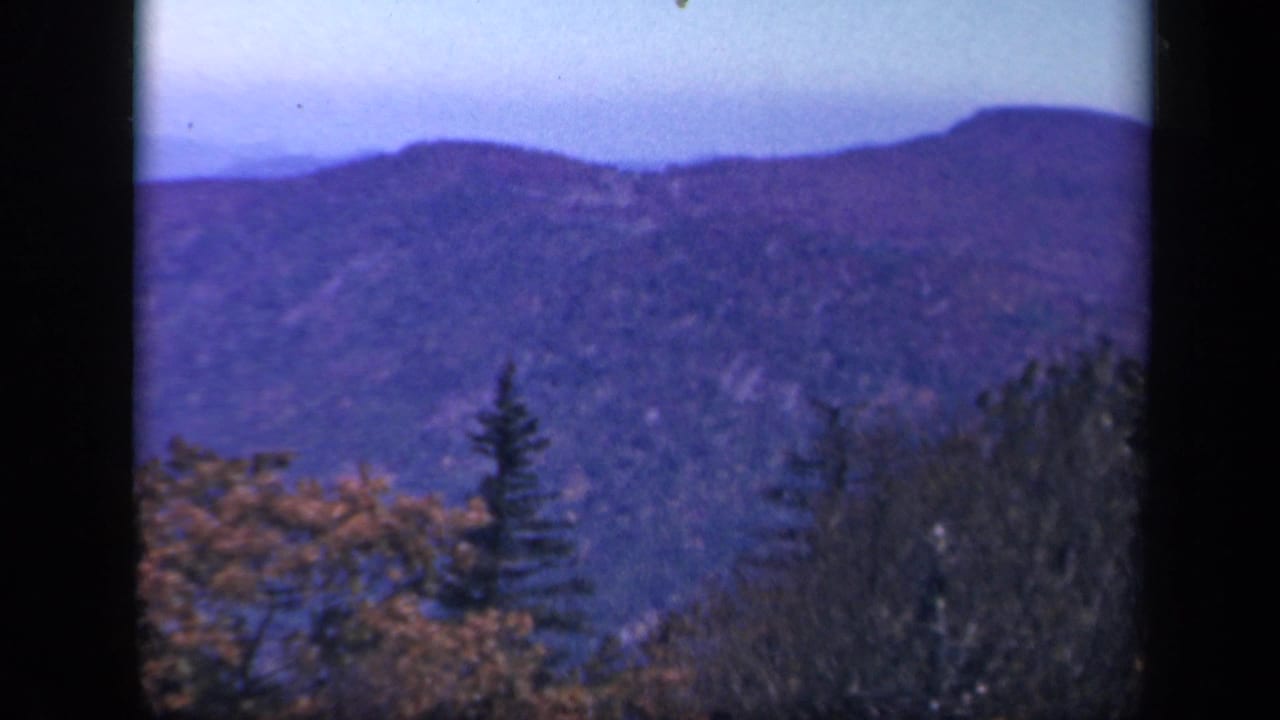 1943 Skyline Drive home-movie frame showing a blue mountain ridge beyond foreground evergreens and fall foliage.