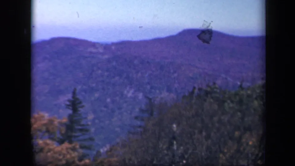1943 8mm Skyline Drive frame with distant ridge, autumn trees, evergreens and a small film mark near the horizon.