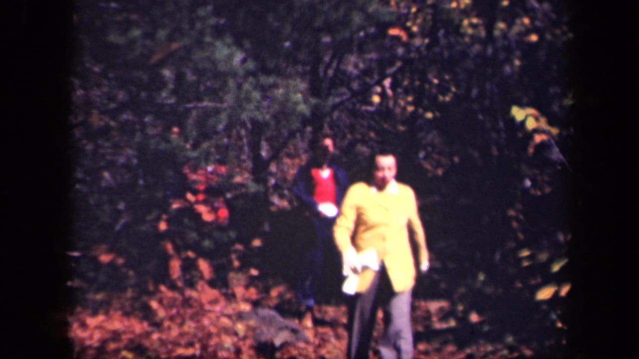 1943 8mm home-movie frame from Skyline Drive showing adults walking through a leaf-covered forest path, led by a person in a yellow jacket.