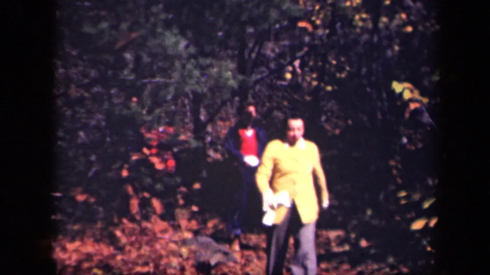1943 8mm home-movie frame from Skyline Drive showing adults walking through a leaf-covered forest path, led by a person in a yellow jacket.