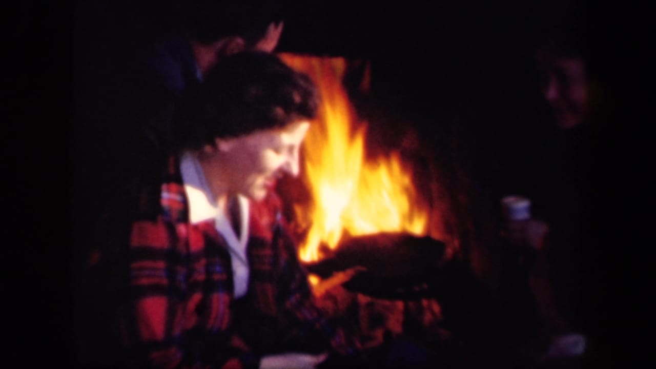 A woman in a red plaid jacket leans beside a campfire while other adults stand or sit nearby.