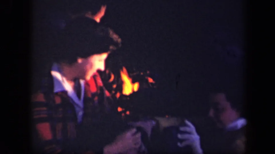 1943 home-movie frame from Skyline Drive showing a plaid-jacketed woman and another seated person beside a small campfire.