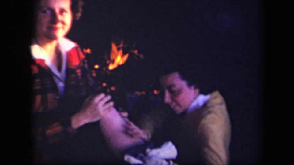 1943 Skyline Drive home-movie frame with a woman in plaid facing camera and another adult seated near the campfire.