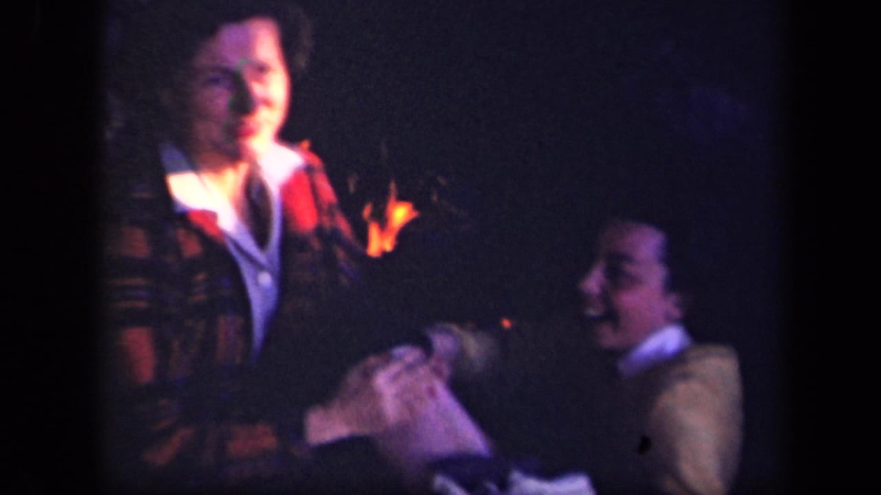 Two people sit near a campfire in dark woods, lit by warm firelight.