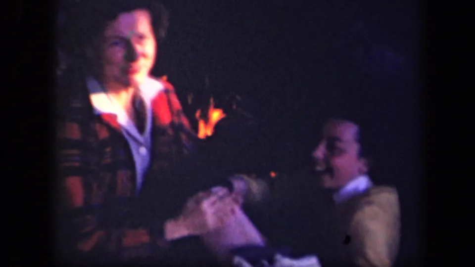 1943 Skyline Drive home-movie frame showing two people near a campfire in dark woods, with soft 8mm color and visible faces.