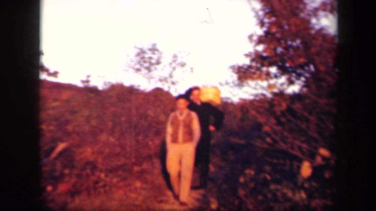 1943 Skyline Drive home-movie frame showing two adult men in a grassy autumn landscape, with one man carrying a large basket behind another.