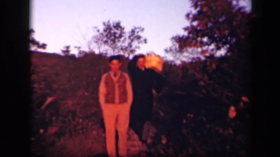 1943 Skyline Drive home-movie frame showing a foreground man facing the camera with another adult man and basket in the brush behind him.