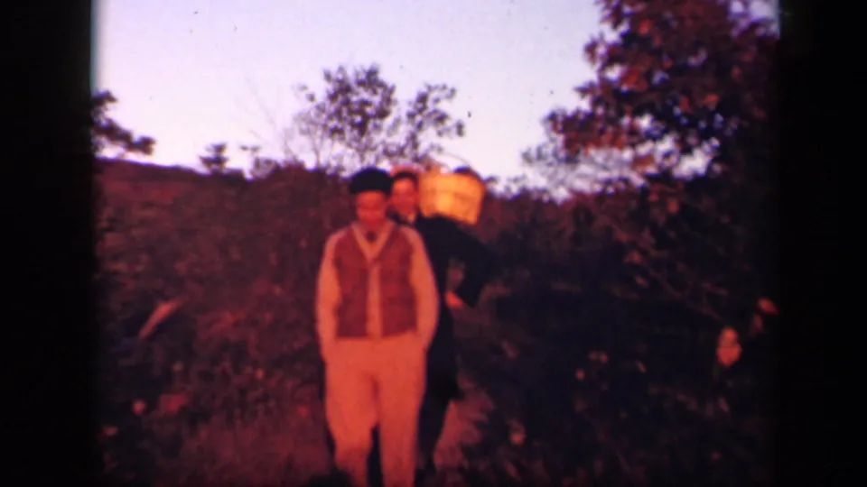 1943 Virginia 8mm frame showing two adult men close together in a dim autumn field, with the large basket still visible behind the front figure.