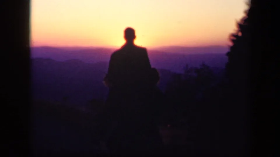 A dark figure remains framed against a purple-orange mountain sunset in archival 8mm footage from Skyline Drive.