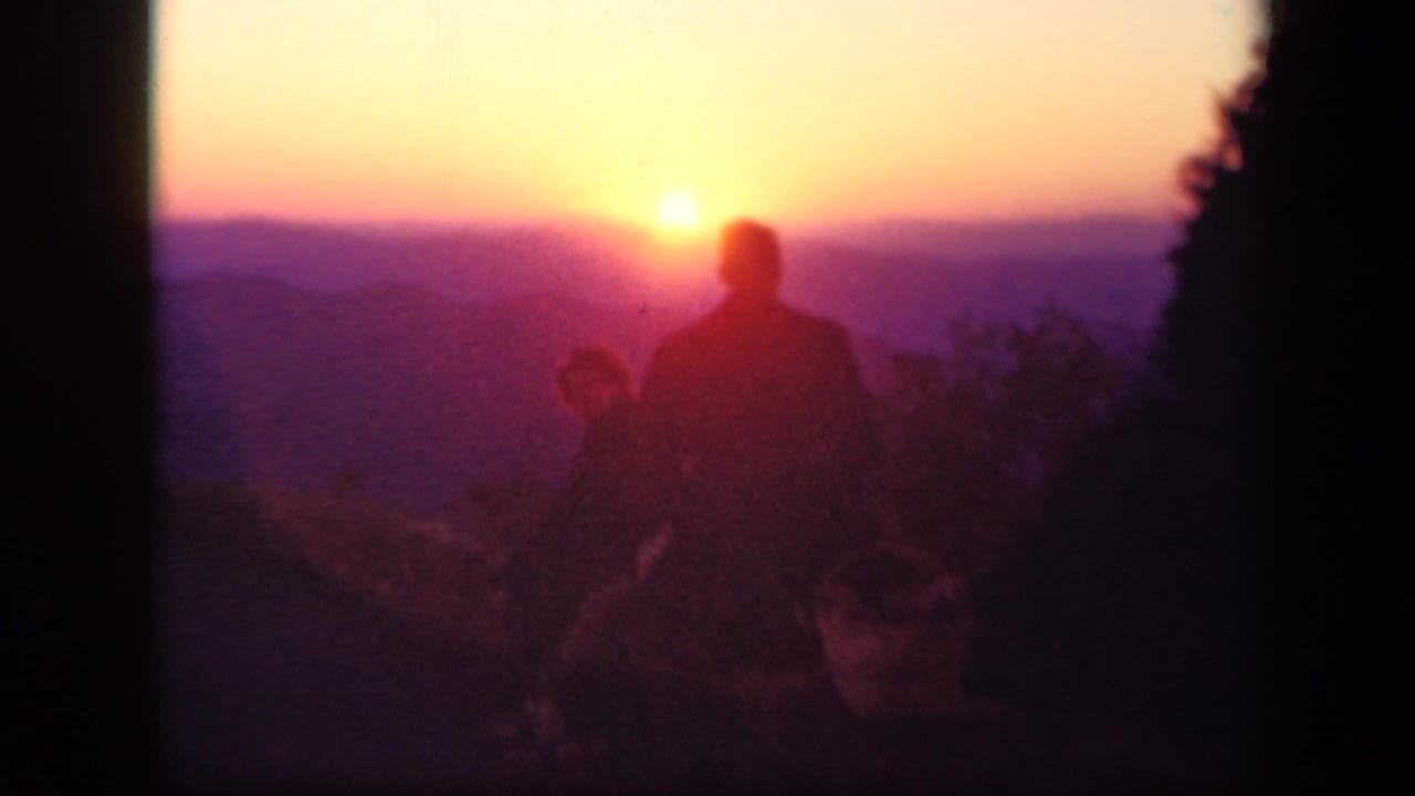 Two dark adult silhouettes sit near a bright sunset over distant hills.