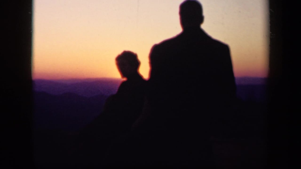 Two silhouetted adults stand before a pink and purple mountain sunset.
