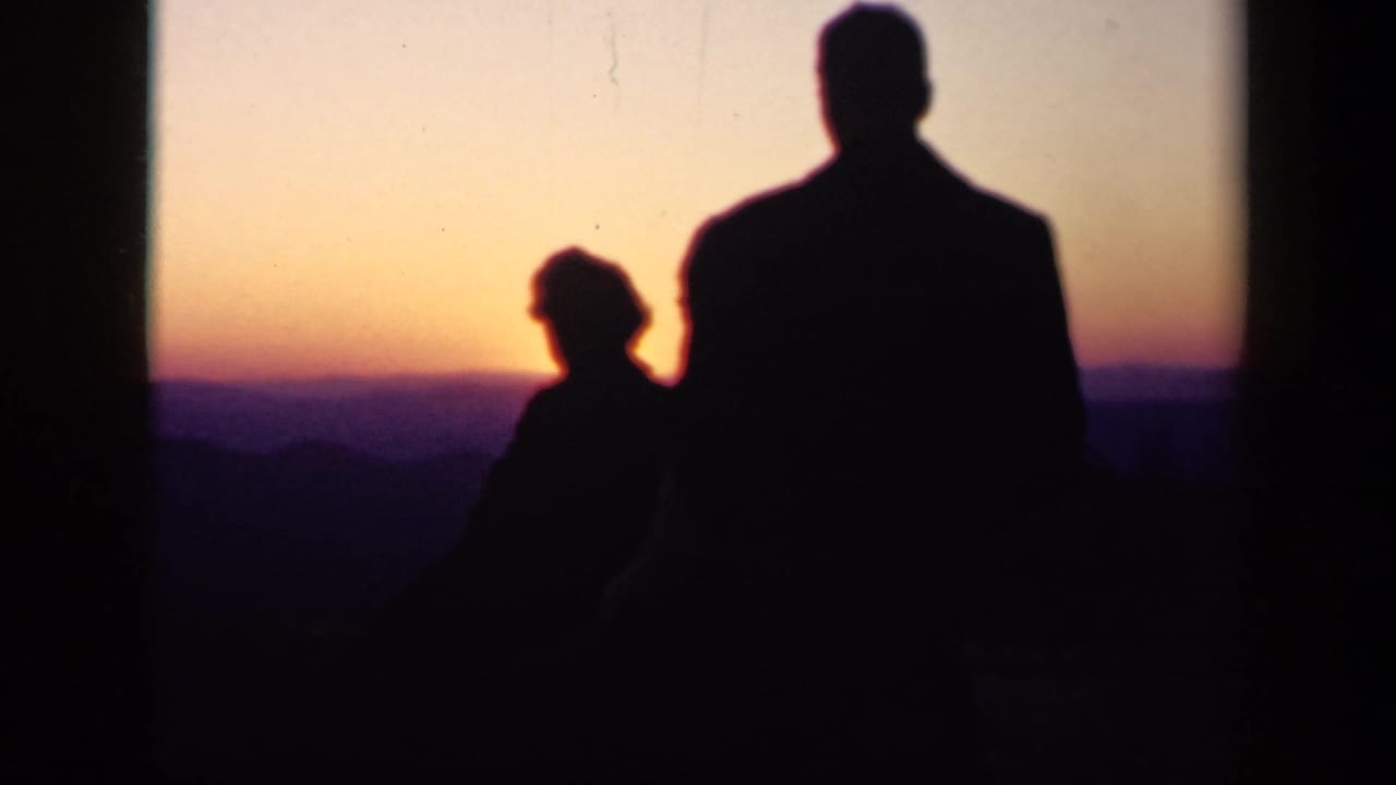 The silhouetted man and woman remain near the overlook edge, looking across Skyline Drive's mountain ridges at sunset.
