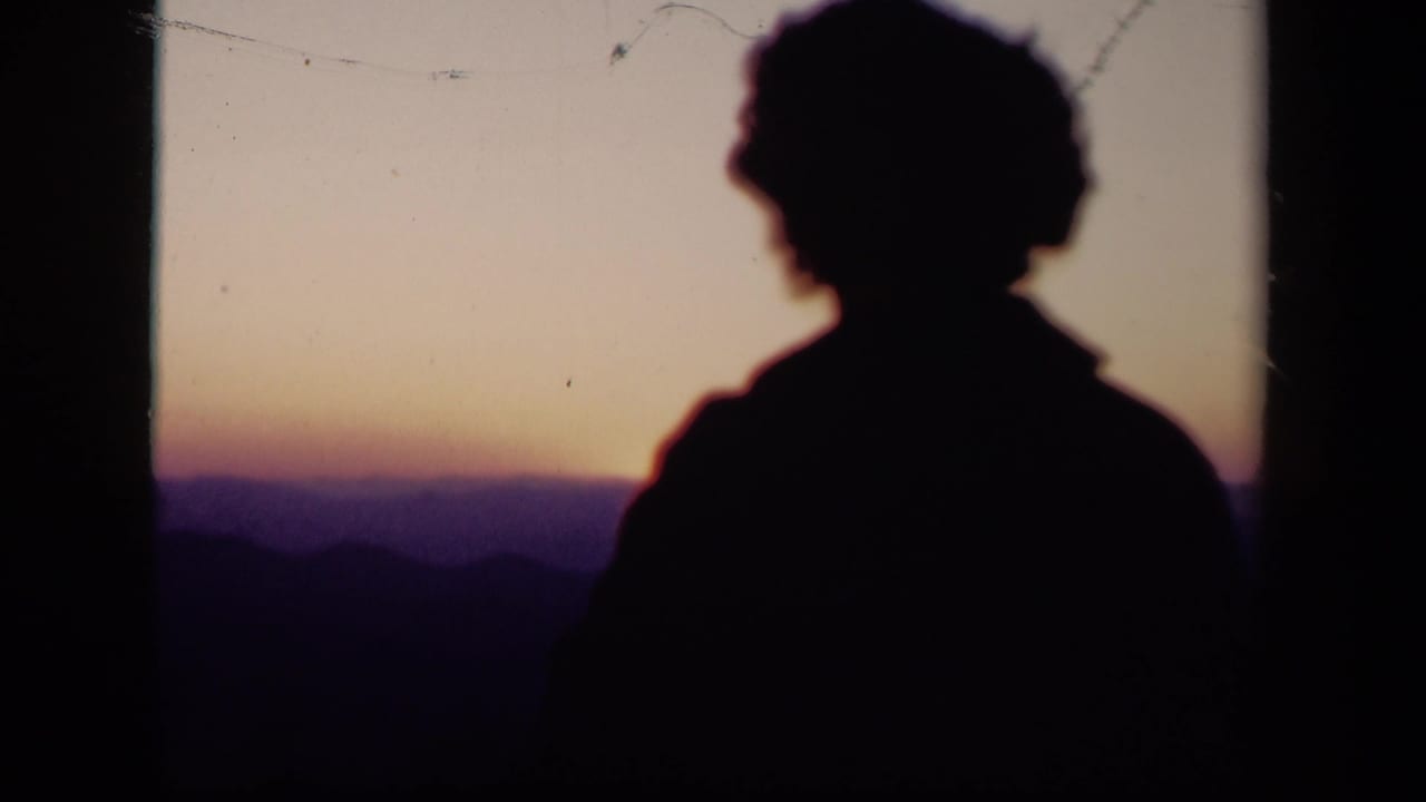 Silhouetted person facing a sunset over distant hills.
