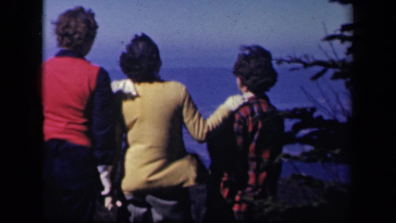 Three women in colorful sweaters stand together at a Skyline Drive overlook, facing a blue distant horizon in 1940s home-movie footage.