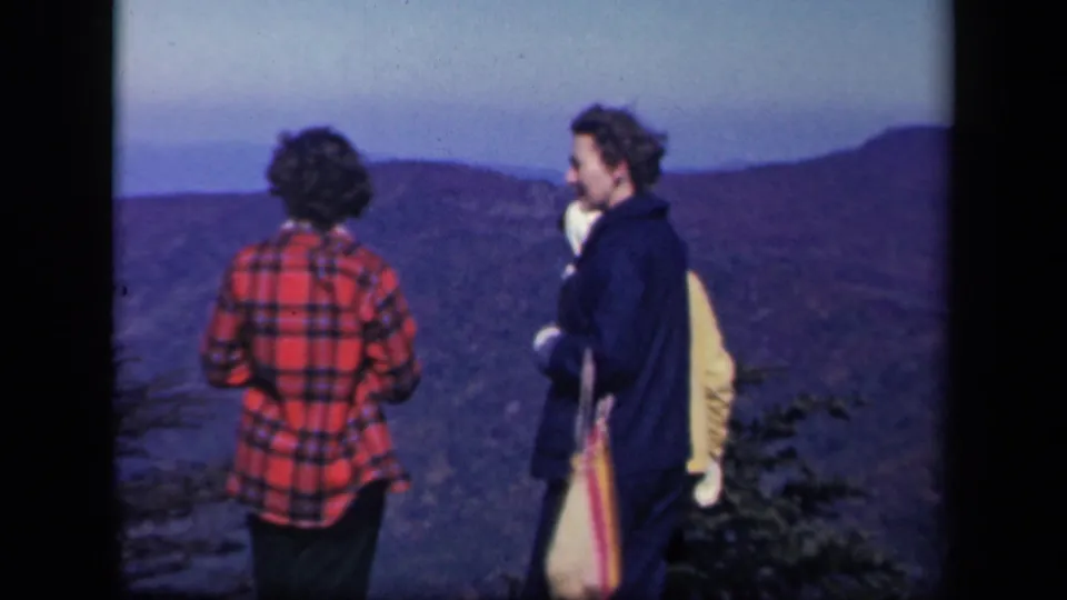 Two women at Skyline Drive turn back toward the camera, with ridges and sky behind them.