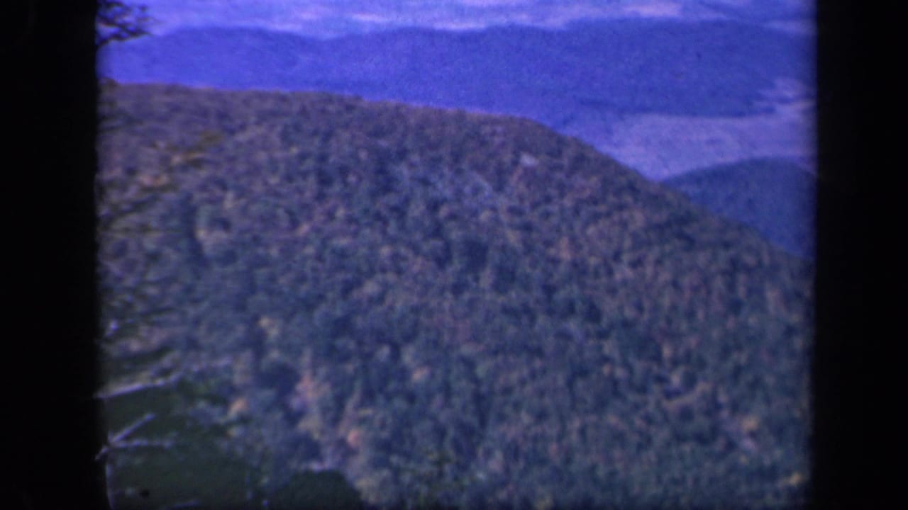 1943 Skyline Drive home-movie frame showing a forested Blue Ridge slope beneath a blue sky and soft distant ridges.