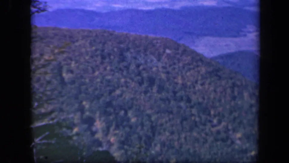 1943 Skyline Drive home-movie frame showing a forested Blue Ridge slope beneath a blue sky and soft distant ridges.