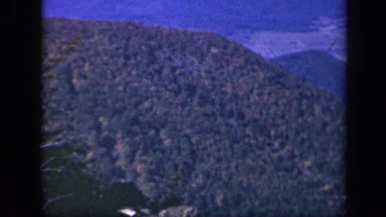 Tree-covered mountain slope with distant ridges and dark projected-film edges.