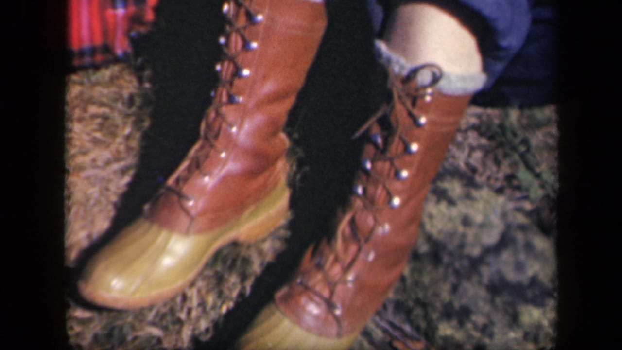 1943 Skyline Drive home-movie frame with brown lace-up boots, yellow toe caps, rolled jeans, and a glimpse of red plaid fabric.