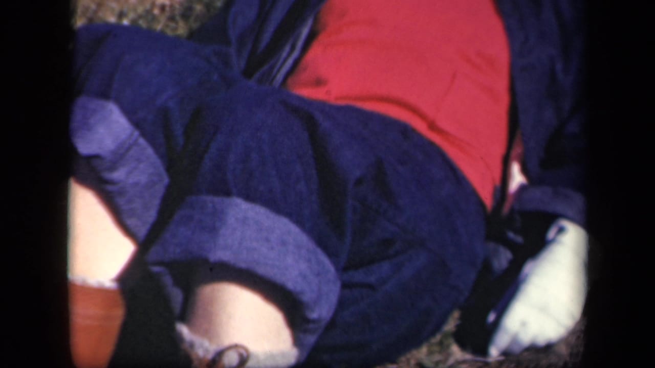 1943 Skyline Drive home-movie frame showing a woman reclining outdoors in a red shirt, dark jacket, and rolled dark trousers.