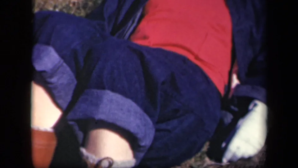 1943 Skyline Drive home-movie frame showing a woman reclining outdoors in a red shirt, dark jacket, and rolled dark trousers.