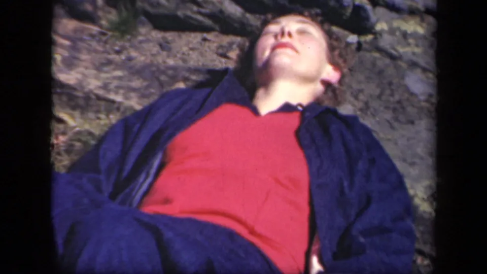 1943 home-movie frame of a woman resting on sunlit ground at Skyline Drive, her face turned upward and eyes closed.