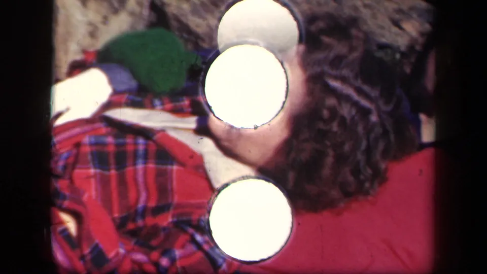 Two women rest beside each other outdoors in the 1943 Skyline Drive footage, with plaid fabric and green gloves visible near bright frame artifacts.