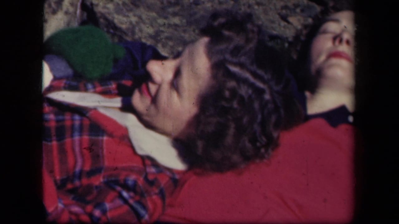 Two adult women lie close together on rocky ground, one turned partly away in a red top.