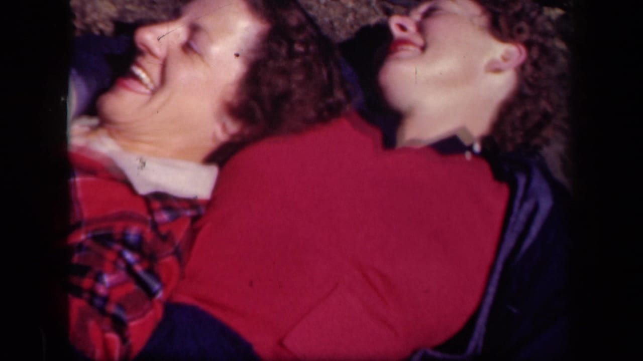 Two women lie close together laughing, one in a red sweater and one in plaid clothing.