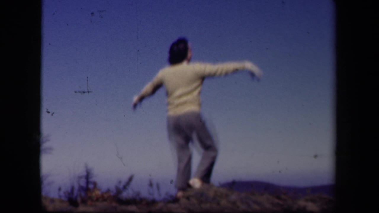 A figure dances with arms out on a rocky Skyline Drive hilltop, with blue sky and distant landscape behind them.