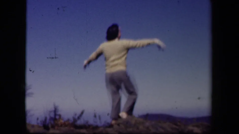 A figure dances with arms out on a rocky Skyline Drive hilltop, with blue sky and distant landscape behind them.