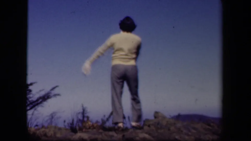A rear view shows the dancer pausing on the Skyline Drive hilltop, with rocky ground, low brush, and blue sky visible.