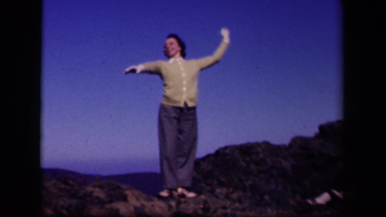 In 1943 Skyline Drive home-movie footage, a woman in a yellow cardigan poses on a rocky hilltop with distant hills behind her.