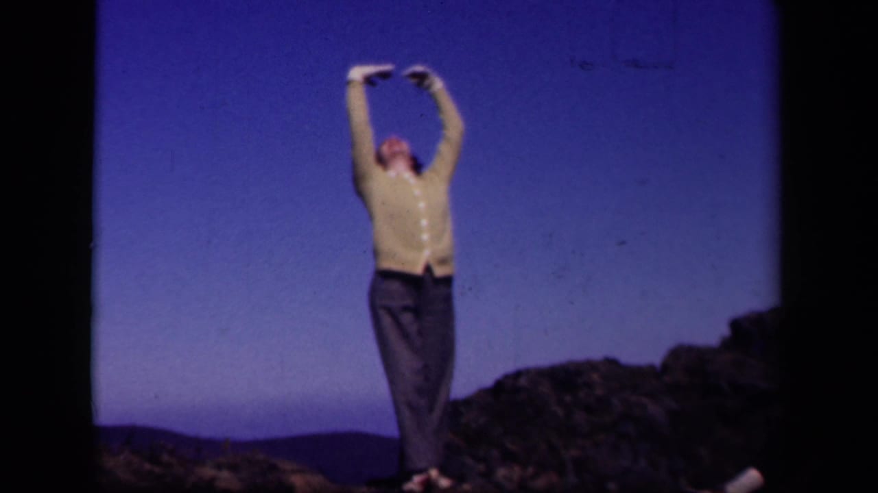 Woman on a rocky hilltop raising both arms overhead beneath a blue sky.