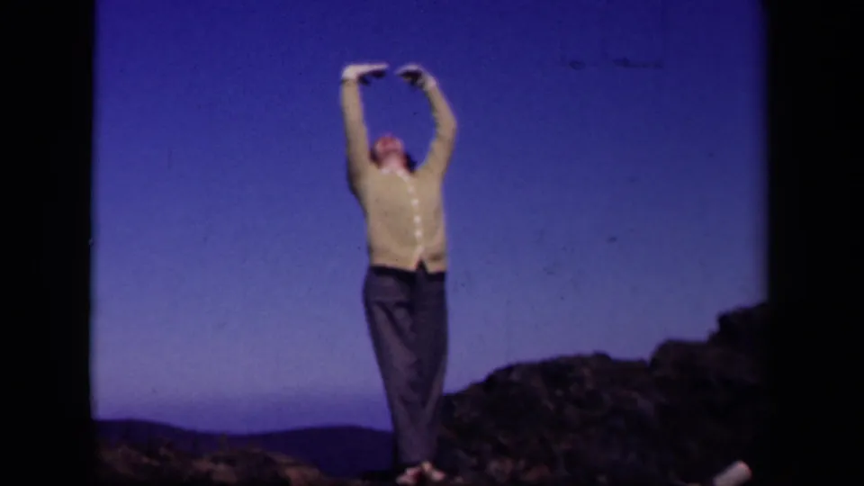 The woman stands on Skyline Drive terrain in the 1943 8mm clip, lifting both arms above her head against the open sky.