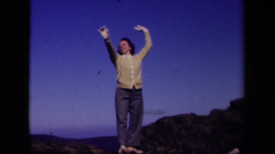 A woman in a yellow cardigan smiles and gestures upward on a Skyline Drive hilltop in this 1943 8mm home-movie frame.