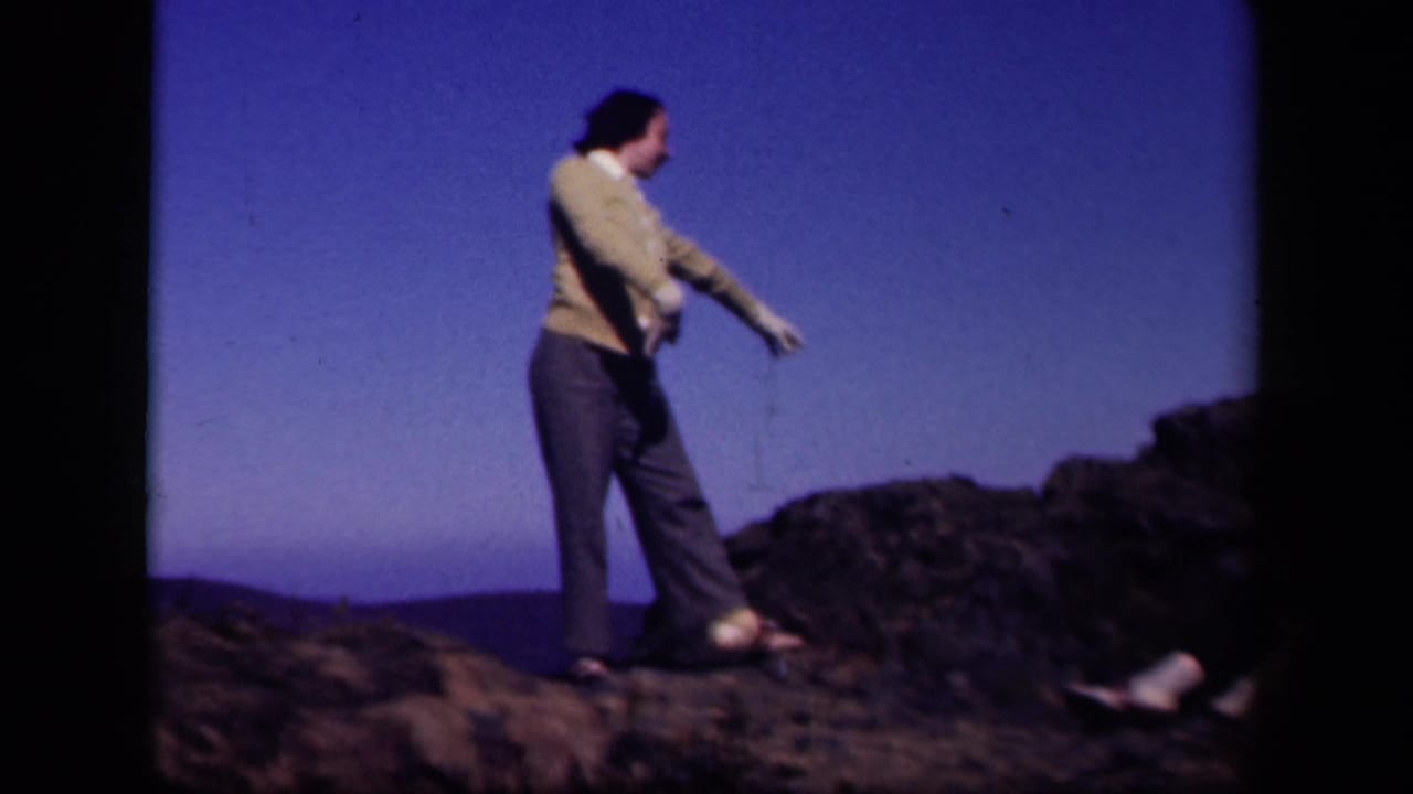 The woman dances sideways on rocks at the Skyline Drive location, with a dark rocky ridge and blue sky behind her.