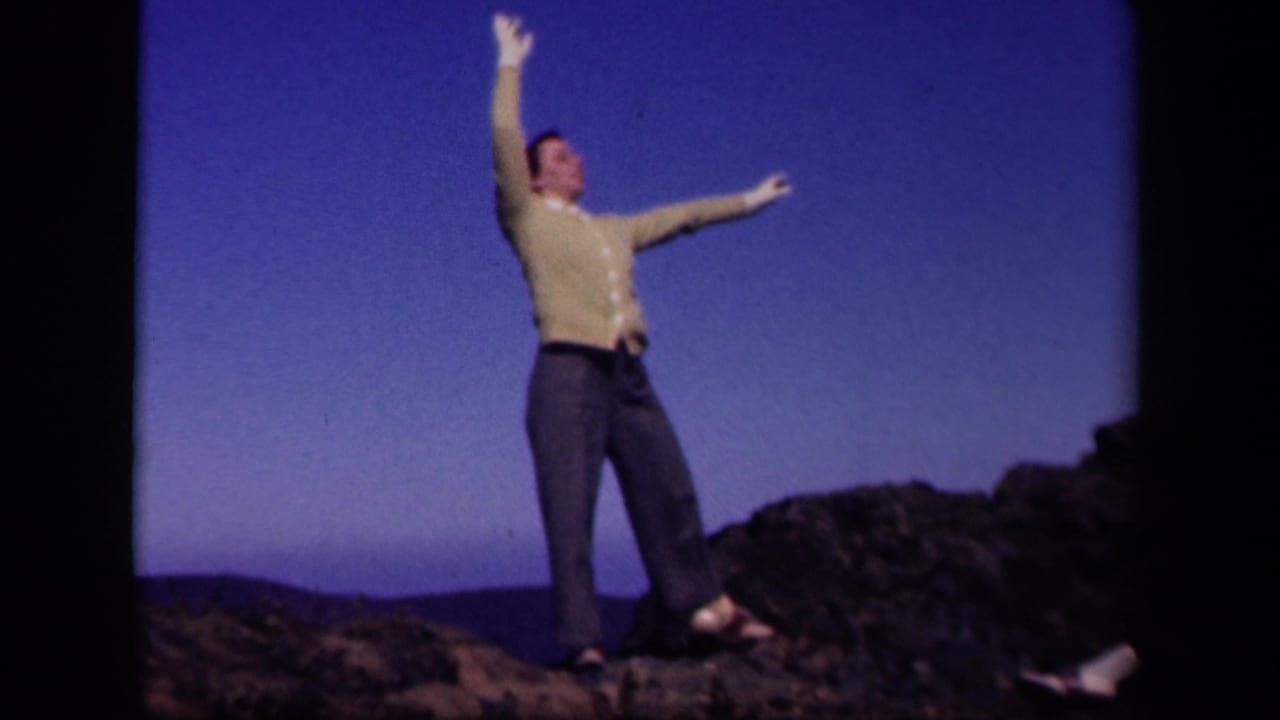 Woman standing on a rocky ridge with one arm raised under a blue sky.