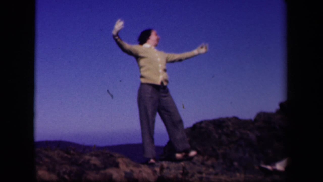 The woman turns with arms lifted on a rocky Skyline Drive overlook, with distant hills behind her.