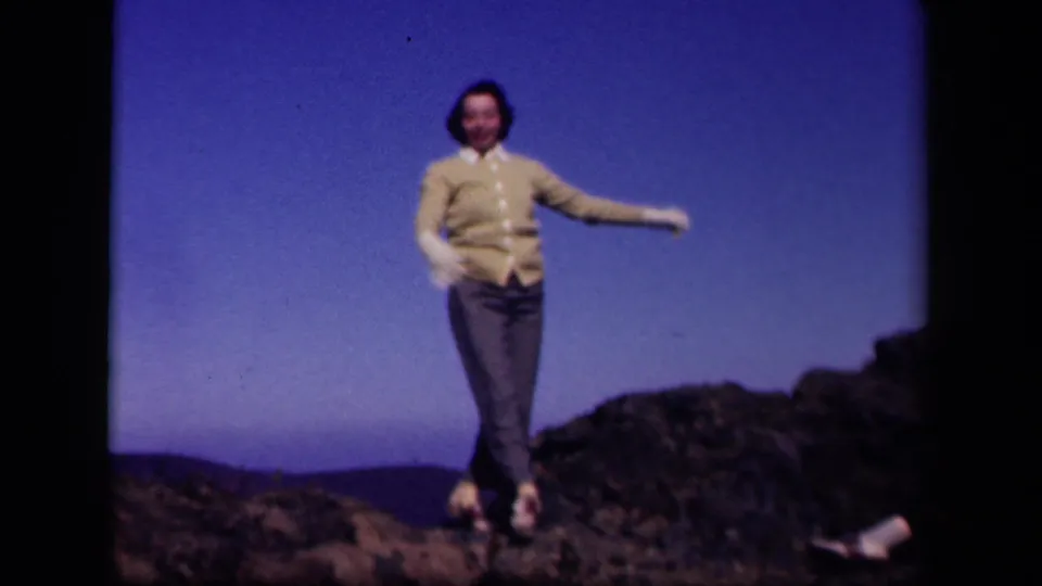 The woman steps forward with one arm extended, framed by rocks and blue sky in the 1943 Skyline Drive home movie.