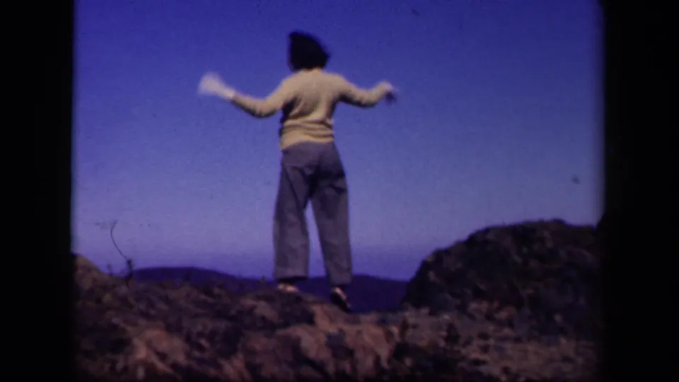 The woman stands on a rocky Skyline Drive overlook with arms spread, framed by blue sky and distant hills.