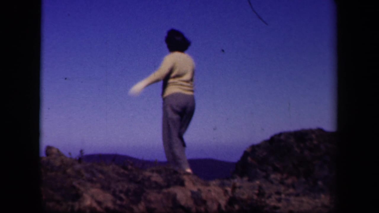 Woman in sweater and gloves steps across rocks under an open blue sky.