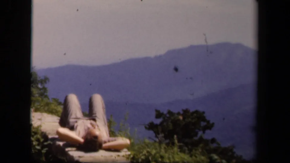 A 1943 Skyline Drive home-movie frame shows a person sunning on a stone wall above distant mountain ridges.