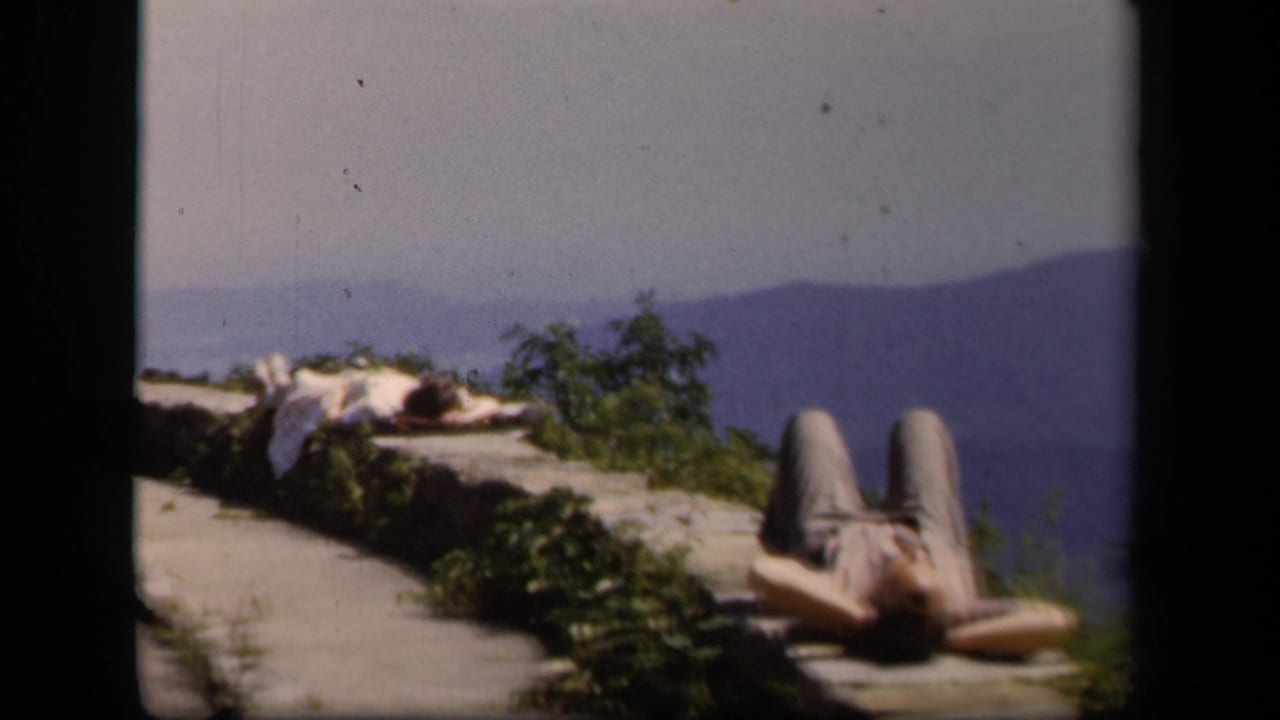 Reclining person on an overlook beside greenery with blue mountains in the background.