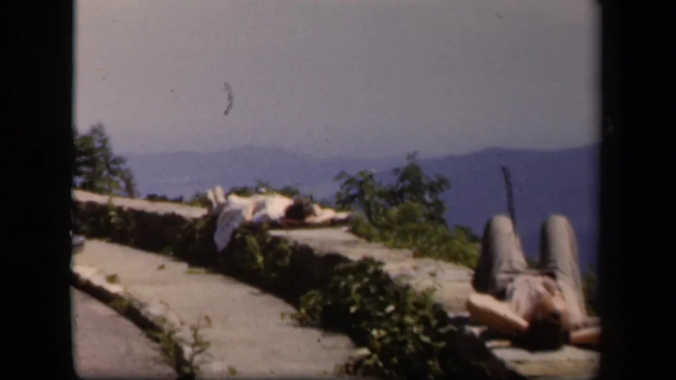 Home-movie view of a Skyline Drive overlook with a person resting on the ledge and mountain scenery behind.