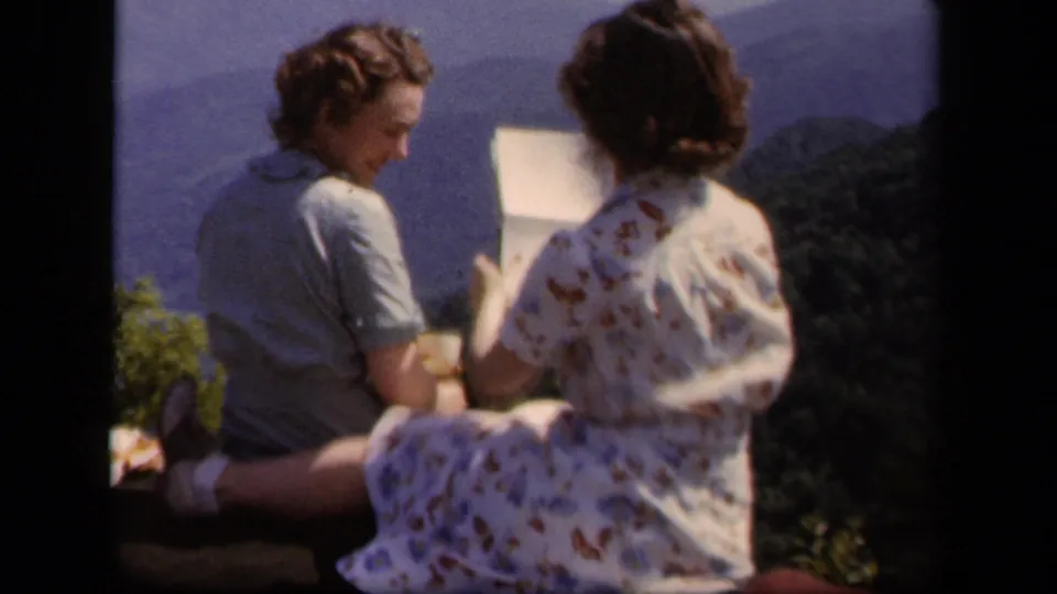 1943 Skyline Drive frame showing two women seated at an overlook, one appearing to read or show a letter, with mountains behind them.