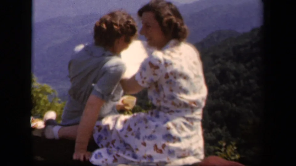 Two women at Skyline Drive turn toward one another beside a paper map, with sunlit mountains and forested slopes behind them.